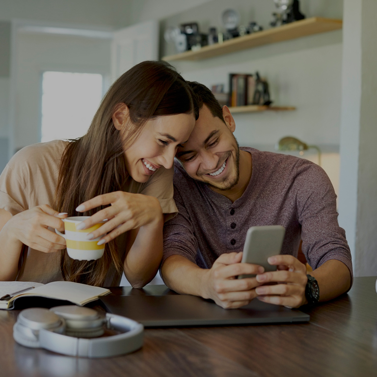 Cheerful boyfriend showing mobile phone to girlfriend in dining room. Young woman is holding coffee cup. They are spending leisure time together at home.