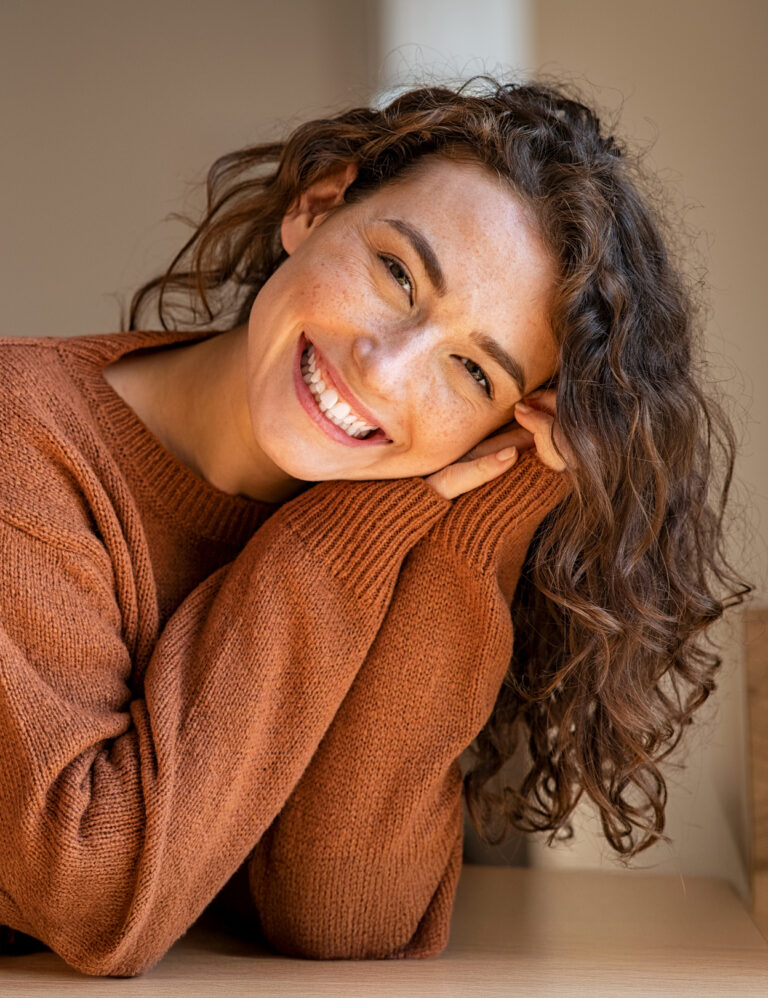 Happy young woman sitting on sofa at home and looking at camera. Portrait of comfortable woman in winter clothes relaxing on a table. Portrait of beautiful girl smiling and relaxing during autumn.